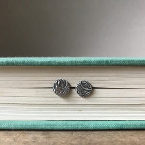 May include: A pair of silver stud earrings with a decorative, swirling pattern. The earrings are shown on a book.