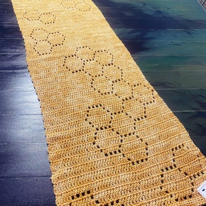 May include: A crocheted table runner in a light brown color with a repeating pattern of circles. The runner is laid out on a dark blue wooden table.