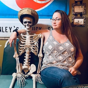 Könnte beinhalten: Ein graues Häkeltop mit einem Totenkopfmuster. Das Top hat einen V-Ausschnitt und wird mit einer blauen Jeans kombiniert. Ein Skelett mit einem beigen Hut sitzt neben der Person, die das Top trägt.