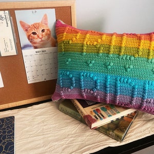 May include: A rainbow colored crocheted pillow with a heart pattern. The pillow is sitting on top of a stack of books.