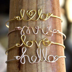 May include: Four metal wire bracelets stacked vertically. The top bracelet is gold-toned with a heart shape, the second is rose gold-toned with the word "mrs", the third is gold-toned with the word "love", and the bottom is silver-toned with "hello".