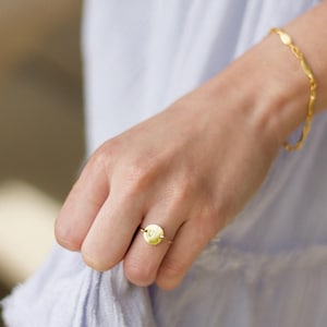 May include: A gold ring featuring a small, circular flat disc charm and a matching thin, wavy gold bracelet. The ring is worn on a finger, and the bracelet is worn on the wrist, both displaying a polished, metallic gold finish throughout.