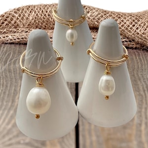 May include: Three gold rings with a white pearl dangling from each. The rings are displayed on white cone-shaped ring holders. The rings are on a wooden surface with a brown burlap fabric in the background.