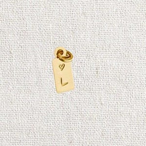 May include: A small, gold-colored rectangular tag with a heart and the letter "L" engraved on it. The tag is suspended from a small loop. The text "L.A. OSBORN" is at the top. The text "14k gold filled personalized mini tags hand-stamped & made to order" is at the bottom.