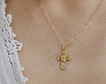 May include: A gold-toned necklace featuring a delicate chain with oval links and a small, textured cross pendant. The cross has a detailed, ornate surface pattern and hangs centrally from the chain, resting against a white textured garment.