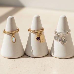 May include: Three rings displayed on white cone stands. The first is a gold ring with an oval opal charm. The second is a gold ring with a purple stone charm and a rectangular tag. The third is a silver ring with a round charm, a clear stone, and a star.