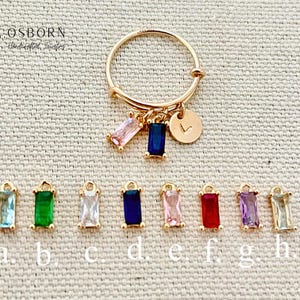 May include: A gold ring with a small heart charm and eight different colored charms with rectangular stones. The charms are labeled a through h.