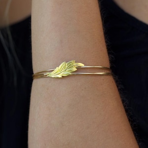 May include: A gold-toned bangle bracelet featuring a central leaf-shaped charm. The bracelet consists of two thin, smooth metallic bands that meet at the detailed, textured leaf element, creating a delicate and layered appearance on the wrist.