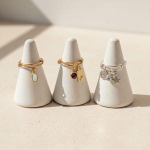 May include: Three rings displayed on white conical stands. The first is a gold ring with an oval opal charm. The second is a gold ring with a purple circular stone and a rectangular tag. The third is a silver ring with a circular charm, a clear stone, and a star.