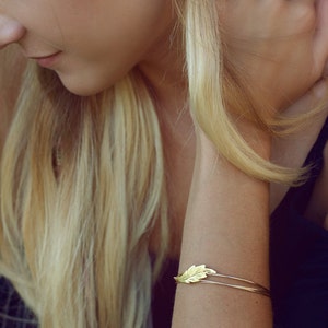 May include: A gold-toned metal bangle bracelet featuring a delicate, textured leaf-shaped charm attached to a thin, double-wire band. The leaf design displays fine, etched vein details across its surface, creating a simple and refined accessory.