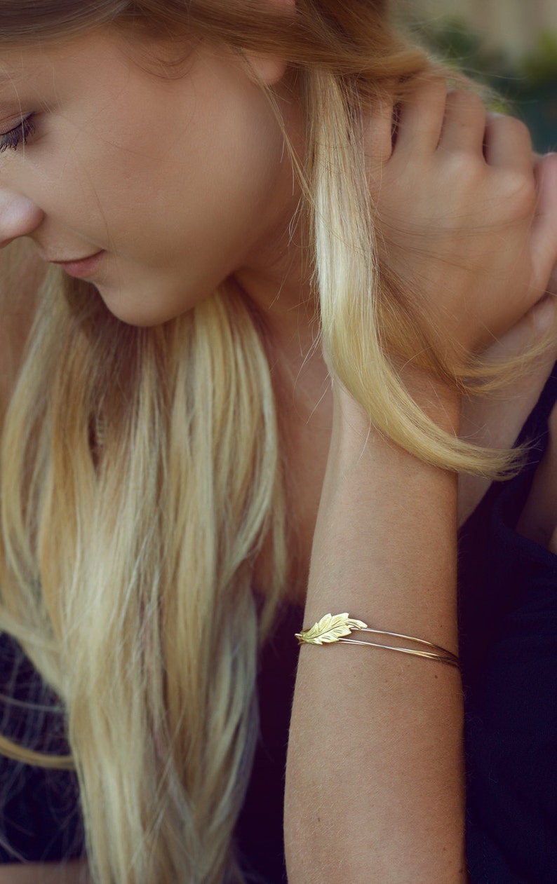 May include: A gold-toned metal bangle bracelet featuring a delicate, textured leaf-shaped charm attached to a thin, double-wire band. The leaf design displays fine, etched vein details across its surface, creating a simple and refined accessory.