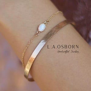 May include: A gold-toned chain bracelet featuring a small, oval-shaped white opal-like stone and a wider, smooth, hammered-finish gold-toned cuff bracelet. The text L.A.OSBORN Handcrafted Jewelry is visible in the center of the image.