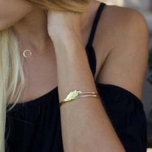 May include: A gold-toned metal cuff bracelet featuring a delicate leaf-shaped design element on the front. The bracelet consists of two thin, parallel bands that meet at the leaf motif. A thin gold-toned chain necklace with a circular pendant is visible.