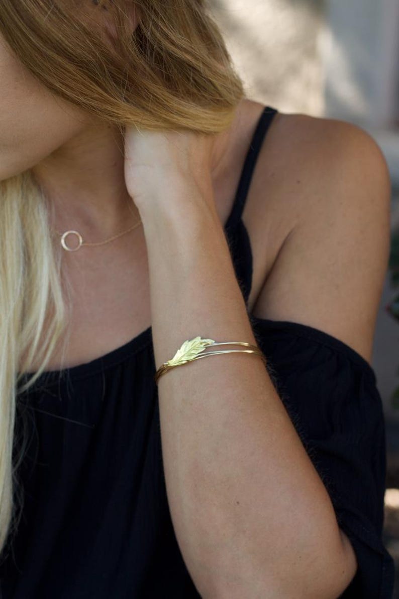 May include: A gold-toned metal cuff bracelet featuring a delicate leaf-shaped design element on the front. The bracelet consists of two thin, parallel bands that meet at the leaf motif. A thin gold-toned chain necklace with a circular pendant is visible.