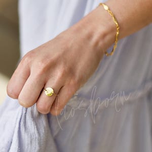 May include: A gold-colored ring with a circular design and a single initial, worn on a finger. A matching gold bracelet is also visible. The jewelry is set against a soft, light blue fabric background.