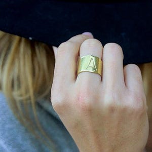 May include: A wide gold-toned band ring featuring a smooth, polished surface with the capital letter A engraved on the front. The ring has a flat, uniform width that encircles the finger, showcasing a metallic finish and a simple, clean design.