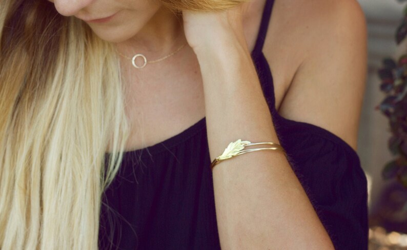 May include: A gold-toned metal cuff bracelet featuring a delicate leaf-shaped design at the center. The bracelet has a thin, open-ended band structure. Additionally, a thin gold-toned chain necklace with a small circular pendant is visible.