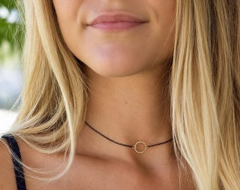 May include: A delicate necklace featuring a thin, dark black chain and a small, textured gold-toned circular pendant centered on the chain. The necklace rests closely against the neck, showcasing a simple design with a contrast between the dark chain and the metallic gold circle.