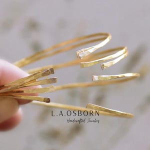 May include: A set of four gold hammered bangle bracelets. The bracelets are stacked on top of each other and are made of thin, flat metal. L.A. OSBORN Handcrafted Jewelry is printed on the bottom of the image.