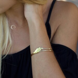 May include: A gold-toned metal cuff bracelet featuring a delicate, textured leaf-shaped centerpiece. The bracelet consists of two thin, parallel bands that meet at the leaf design, creating a sleek and open-ended structure on the wrist.