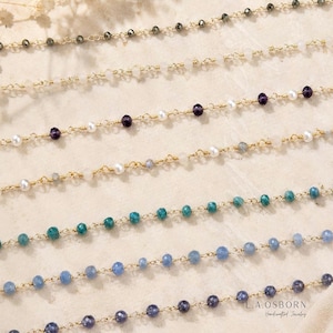 May include: A collection of seven delicate gold-toned wire-wrapped chains featuring various gemstone beads, including dark gray, white, purple, teal, and blue hues. The text L.A. OSBORN Handcrafted Jewelry is printed in the bottom right corner.