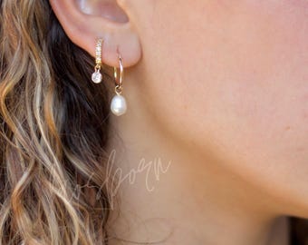May include: Two gold hoop earrings worn in the earlobe. One hoop features a small, clear, round-cut stone charm, while the other hoop features a single, white, teardrop-shaped pearl pendant hanging from the bottom of the thin gold metal ring.
