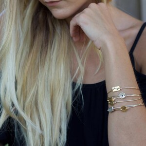 May include: A stack of five thin metal bangles worn on the wrist. Each bangle features a small, distinct charm, including rectangular and circular shapes in gold and silver tones. The bracelets are delicate, wire-like, and sit together on the arm.