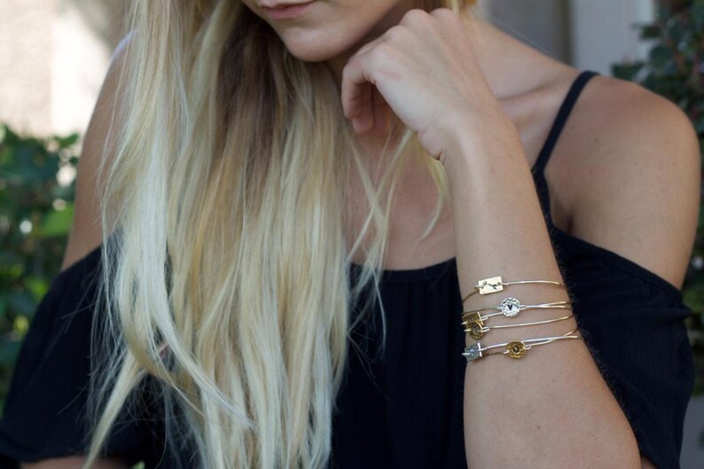 May include: A stack of five thin metal bangles worn on the wrist. Each bangle features a small, distinct charm, including rectangular and circular shapes in gold and silver tones. The bracelets are delicate, wire-like, and sit together on the arm.