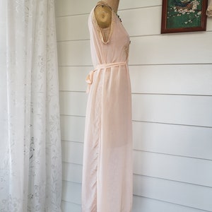 Puede incluir: Camis&oacute;n vintage rosa p&aacute;lido exhibido en un maniqu&iacute;. El camis&oacute;n presenta delicados adornos de encaje a lo largo del escote y el dobladillo, con un cintur&oacute;n a juego en la cintura. La tela parece ligera y fluida.