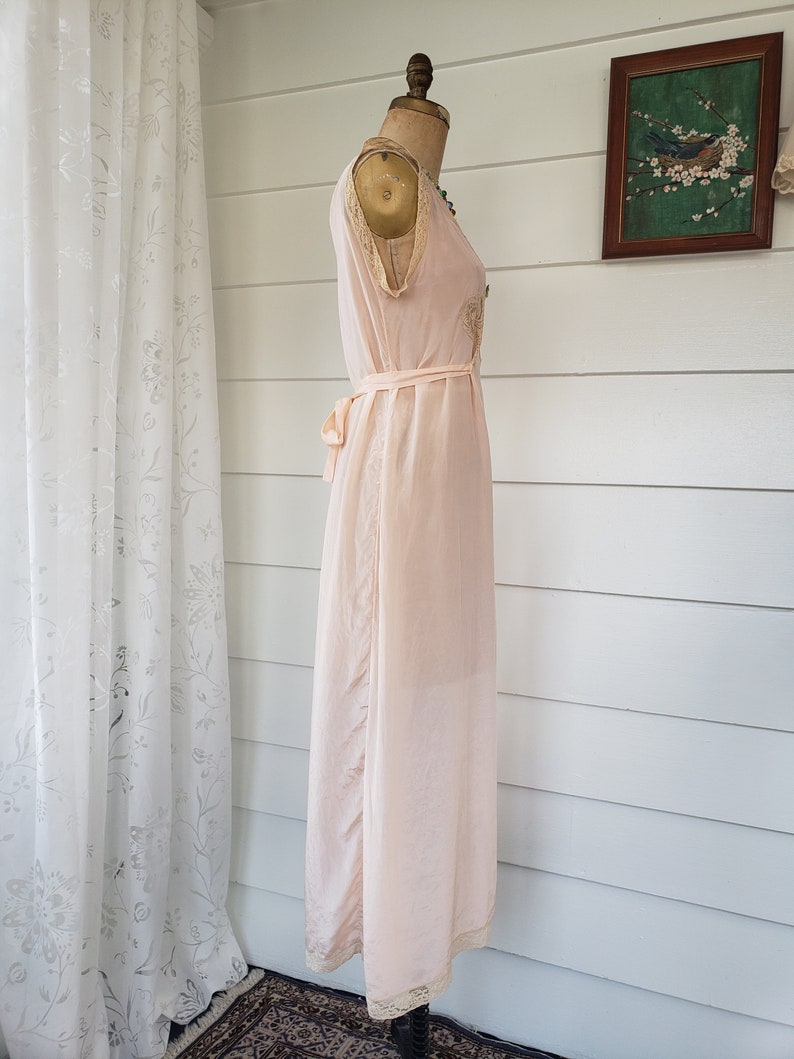 Puede incluir: Camis&oacute;n vintage rosa p&aacute;lido exhibido en un maniqu&iacute;. El camis&oacute;n presenta delicados adornos de encaje a lo largo del escote y el dobladillo, con un cintur&oacute;n a juego en la cintura. La tela parece ligera y fluida.