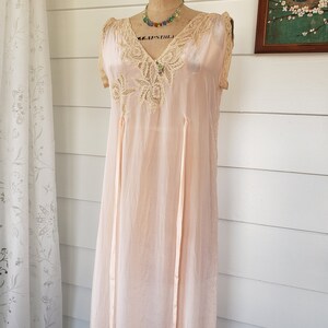 Puede incluir: Un camis&oacute;n vintage de color rosa p&aacute;lido sobre un maniqu&iacute;. El camis&oacute;n presenta un escote en V con detalles de encaje y delicados bordados. Dos cintas largas y finas recorren la parte delantera. Un collar de cuentas de colores y un peque&ntilde;o broche adornan el escote. El vestido se exhibe frente a una pared blanca.