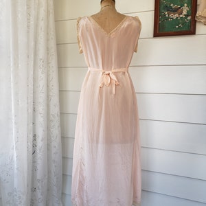Puede incluir: Vestido interior vintage rosa p&aacute;lido sobre un maniqu&iacute;. El vestido presenta un escote en V, delicados adornos de encaje y un cintur&oacute;n a juego en la cintura. El vestido interior es hasta la rodilla y tiene un brillo sutil, posiblemente seda o sat&eacute;n.