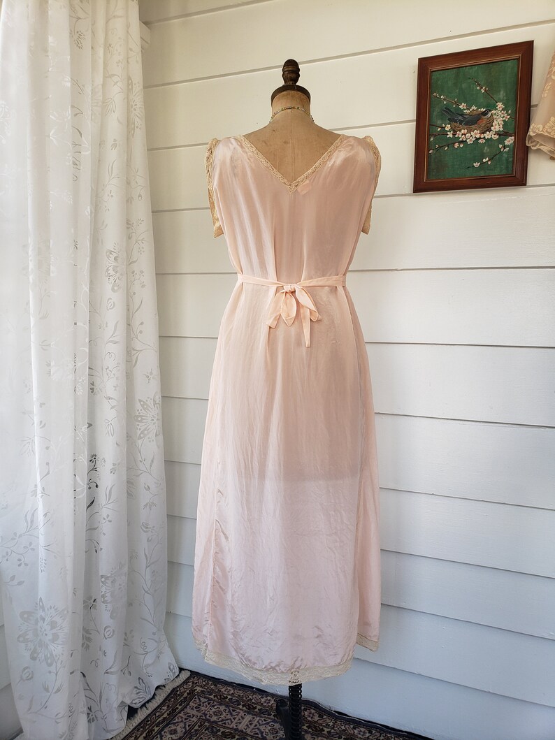 Puede incluir: Vestido interior vintage rosa p&aacute;lido sobre un maniqu&iacute;. El vestido presenta un escote en V, delicados adornos de encaje y un cintur&oacute;n a juego en la cintura. El vestido interior es hasta la rodilla y tiene un brillo sutil, posiblemente seda o sat&eacute;n.