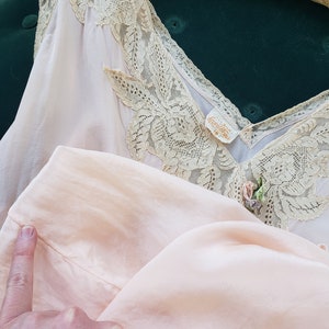 Puede incluir: Conjunto de lencer&iacute;a vintage rosa p&aacute;lido que incluye una camisola y una parte inferior a juego. La camisola tiene un escote en V adornado con delicado encaje. Se puede ver una peque&ntilde;a etiqueta. El conjunto se muestra en una percha sobre un fondo verde oscuro.