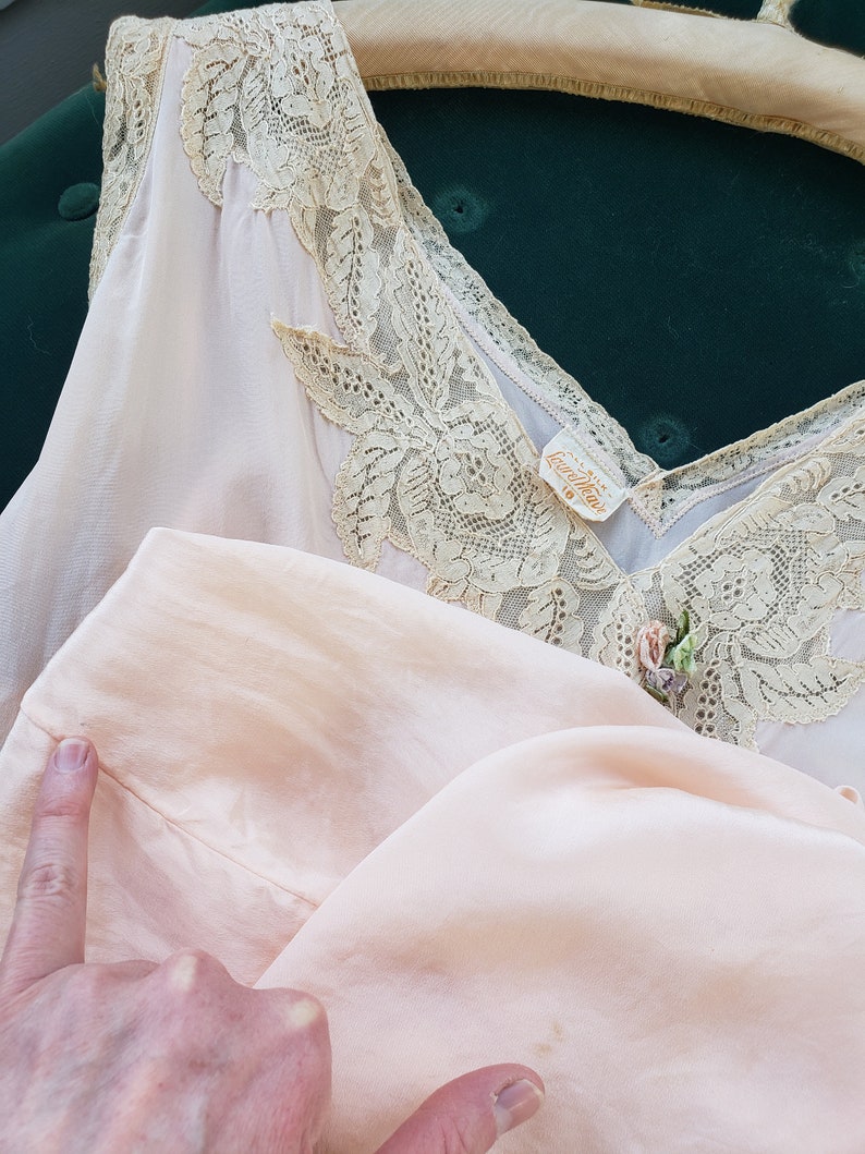 Puede incluir: Conjunto de lencer&iacute;a vintage rosa p&aacute;lido que incluye una camisola y una parte inferior a juego. La camisola tiene un escote en V adornado con delicado encaje. Se puede ver una peque&ntilde;a etiqueta. El conjunto se muestra en una percha sobre un fondo verde oscuro.