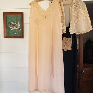 Puede incluir: Un camis&oacute;n vintage color melocot&oacute;n con ribete de encaje y un dise&ntilde;o de mariposa decorativa. Una chaqueta de encaje crema transparente y un vestido negro cuelgan cerca. Se ve una pintura enmarcada de un p&aacute;jaro en una rama.