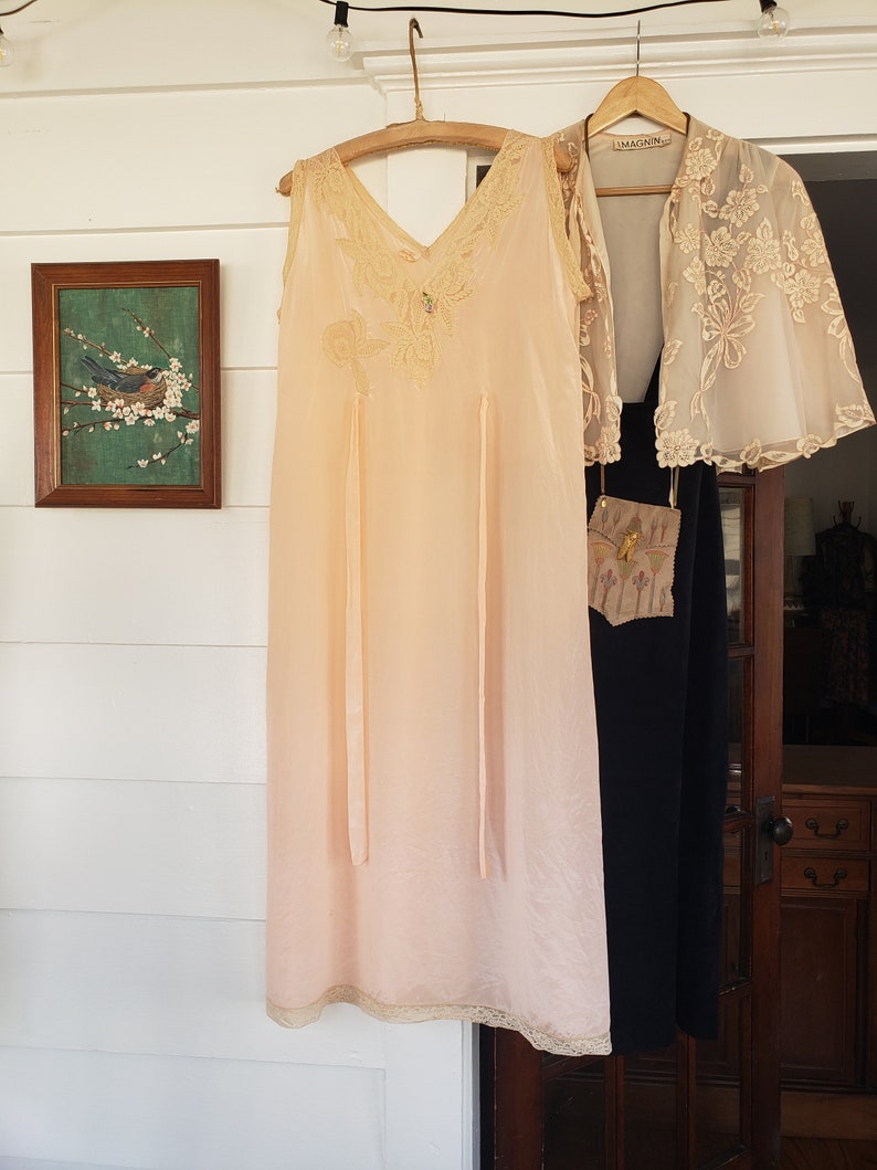 Puede incluir: Un camis&oacute;n vintage color melocot&oacute;n con ribete de encaje y un dise&ntilde;o de mariposa decorativa. Una chaqueta de encaje crema transparente y un vestido negro cuelgan cerca. Se ve una pintura enmarcada de un p&aacute;jaro en una rama.