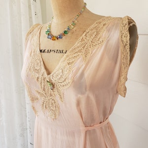 Puede incluir: Un vestido de estilo vintage, rosa p&aacute;lido, con delicados adornos de encaje y un cintur&oacute;n a juego. El vestido presenta un escote en V y detalles de encaje intrincados en el corpi&ntilde;o. Un collar de cuentas de colores con cuentas de varios colores adorna el cuello del maniqu&iacute;.