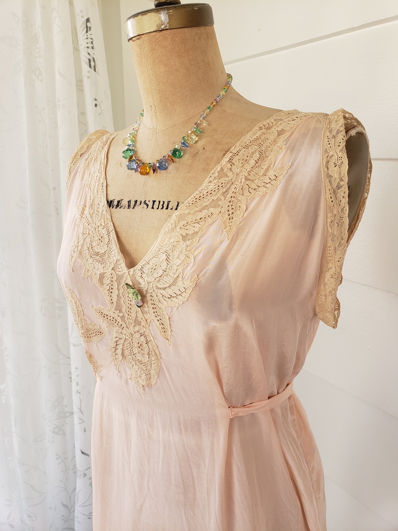 Puede incluir: Un vestido de estilo vintage, rosa p&aacute;lido, con delicados adornos de encaje y un cintur&oacute;n a juego. El vestido presenta un escote en V y detalles de encaje intrincados en el corpi&ntilde;o. Un collar de cuentas de colores con cuentas de varios colores adorna el cuello del maniqu&iacute;.