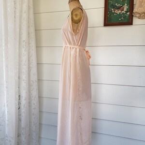 Puede incluir: Camis&oacute;n vintage rosa p&aacute;lido exhibido en un maniqu&iacute;. El camis&oacute;n sin mangas presenta un delicado ribete de encaje a lo largo del escote y el dobladillo, con un cintur&oacute;n de cinta a juego atado a la cintura. La tela parece ligera y fluida.