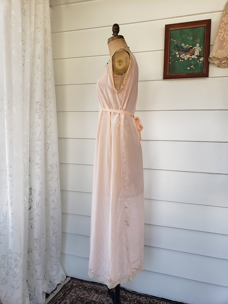 Puede incluir: Camis&oacute;n vintage rosa p&aacute;lido exhibido en un maniqu&iacute;. El camis&oacute;n sin mangas presenta un delicado ribete de encaje a lo largo del escote y el dobladillo, con un cintur&oacute;n de cinta a juego atado a la cintura. La tela parece ligera y fluida.