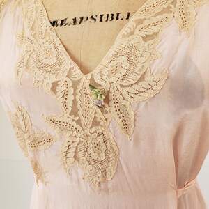 Puede incluir: Vestido vintage rosa p&aacute;lido con intrincados detalles de encaje beige alrededor del cuello en V y las mangas. Un collar de cuentas de colores y un peque&ntilde;o broche floral a&ntilde;aden acentos. El vestido se exhibe en un maniqu&iacute; vintage.