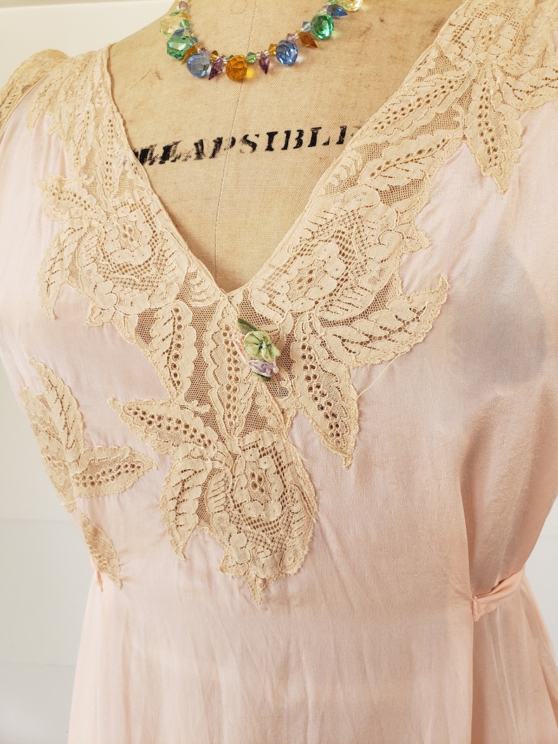 Puede incluir: Vestido vintage rosa p&aacute;lido con intrincados detalles de encaje beige alrededor del cuello en V y las mangas. Un collar de cuentas de colores y un peque&ntilde;o broche floral a&ntilde;aden acentos. El vestido se exhibe en un maniqu&iacute; vintage.