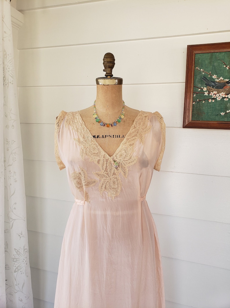 Puede incluir: Un vestido interior rosa p&aacute;lido vintage con detalles de encaje en el escote y los hombros. El vestido se exhibe en un maniqu&iacute; y se combina con un collar de cuentas de colores. Una pintura enmarcada de un p&aacute;jaro es visible en el fondo.