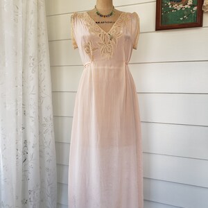 Puede incluir: Un vestido de seda rosa rubor vintage con delicados adornos y detalles de encaje. El vestido presenta un escote en V y mangas cortas, exhibido en un maniqu&iacute;. Un collar de cuentas de colores a&ntilde;ade un toque de color. El vestido es largo y tiene una est&eacute;tica rom&aacute;ntica y femenina.