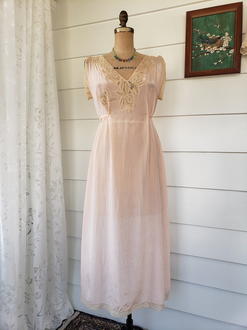 Puede incluir: Un vestido de seda rosa rubor vintage con delicados adornos y detalles de encaje. El vestido presenta un escote en V y mangas cortas, exhibido en un maniqu&iacute;. Un collar de cuentas de colores a&ntilde;ade un toque de color. El vestido es largo y tiene una est&eacute;tica rom&aacute;ntica y femenina.