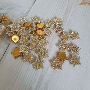 May include: A group of gold-toned star-shaped embellishments with clear iridescent rhinestones. The embellishments have a flat back with a gold-toned metal backing.