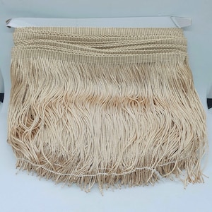 May include: A roll of beige fringe trim. The trim has a woven top section and long, flowing strands of fringe. The fringe is a light, neutral colour, suitable for various craft and design projects. The trim is attached to a white backing.