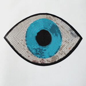 Op de afbeelding: Een decoratieve oogvormige patch met een zwarte rand en een witte achtergrond met zwarte stippen. Het oog heeft een helderblauwe iris en een zwarte pupil. De patch is waarschijnlijk gemaakt van pailletten.