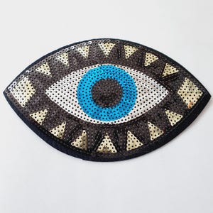 May include: A decorative eye-shaped patch with a black felt base. The eye features a blue iris, a white and black dotted pupil, and a border of black and gold sequins. The patch is designed for embellishment.